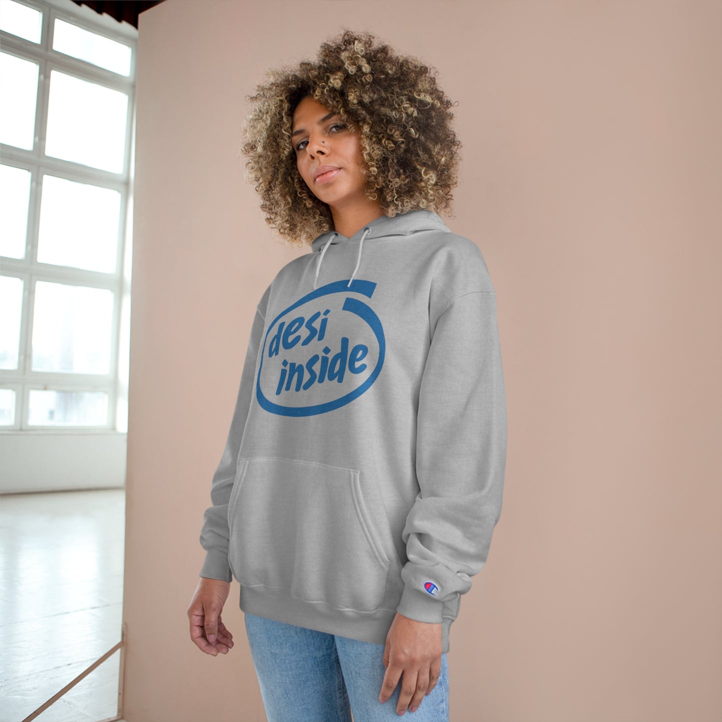 Woman wearing a gray 'Desi Inside' Champion hoodie in a cozy indoor setting.