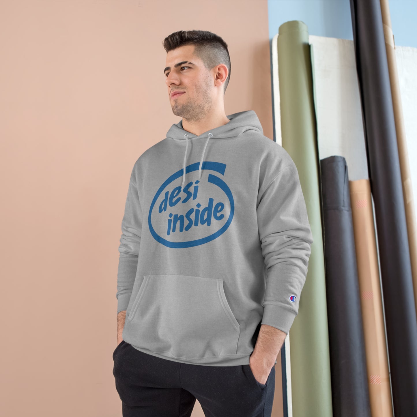 Model wearing a gray Desi Inside Champion hoodie, showcasing the logo and spacious pocket against a colorful backdrop.